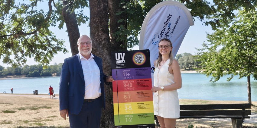 UV-Index-Tafeln Foto: Ein Mann und eine Frau stehen vor einem Baum an einem See und halten eine Tafel mit mehreren Farben - eine UV-Index-Tafel - in die Kamera