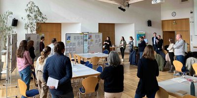 Foto: Teilnehmende eines Workshops stehen im Halbkreis um zwei Infotafeln und diskutieren.