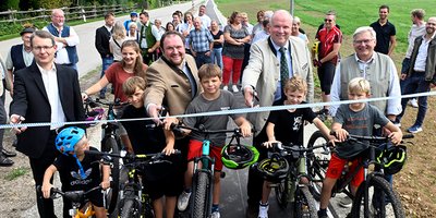 Foto: Mehrere Kinder auf Fahrrädern, dahinter schneiden vier Männer ein Band durch, zahlreiche Zuschauer stehen dahinter.