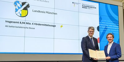 Foto: Im Hintergrund eine Leinwand mit Präsentation, auf der das Landkreiswappen und die Fördersumme zu sehen sind. Rechts im Vordergrund stehen Bundesverkehrsminister Patrick Schnieder und Projektleiter Dominik Scheuer aus dem Landratsamt München. Sie halten beide eine Urkunde mit der Förderzusage in die Kamera.
