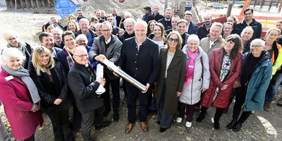Foto: Viele Menschen stehen vor einer Baustelle. Drei von ihnen halten eine Zeitkapsel und die dazugehörigen Unterlagen in die Kamera.