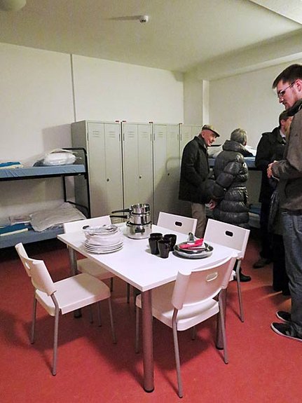 Foto: Besucher besichtigen ein Zimmer der Asylbewerberunterkunft. Im Zimmer befinden sich Stockbetten, Spinde sowie ein Tisch mit Stühlen. Auf dem Tisch steht Geschirr für die Asylbewerber bereit.