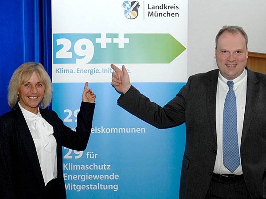 Foto: Landrat Christoph Göbel und Haars Erste Bürgermeisterin Gabriele Müller stehen von einem Roll-Up zur Kampagne und zeigen mit ihren Fingern auf das Logo der Kampagne 29++ Klima. Energie. Initiative.