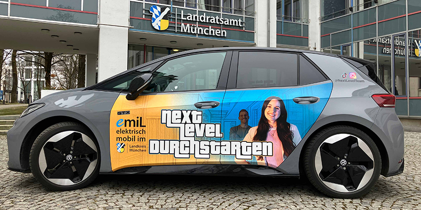Ein Auto, VW id3, mit Folierung im Stil der neuen Arbeitgebermarke des Landkreises steht seitlich vor dem Haupteingang des Landratsamts.